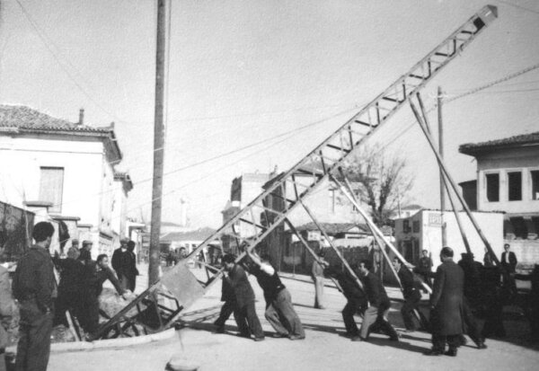 Προσπάθεια εργατών της Υπηρεσίας Ηλεκτροφωτισμού του Δήμου Λάρισας (ΟΥΗΛ) να ανυψώσουν σιδερένιο στύλο, στη διασταύρωση των οδών 23ης Οκτωβρίου & Λ. Κατσώνη. Χρονολογία: μέσα της δεκαετίας του 1950. Φωτογράφος: Τάκης Τλούπας.